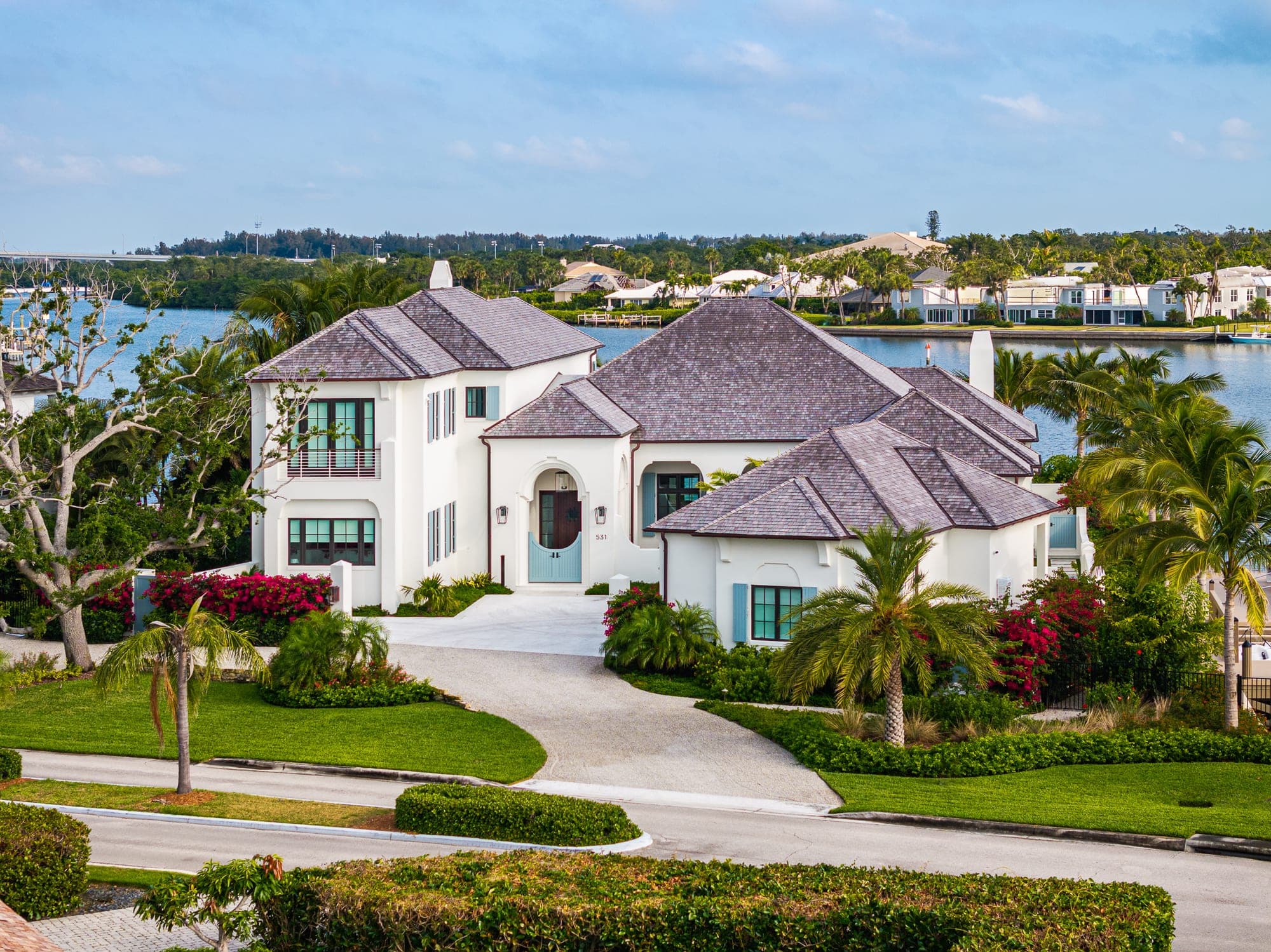 Aerial view of the front of a white stucco riverfront home with a gray shingle roof, an arched blue entry door and tropical landscaping, Vero Beach