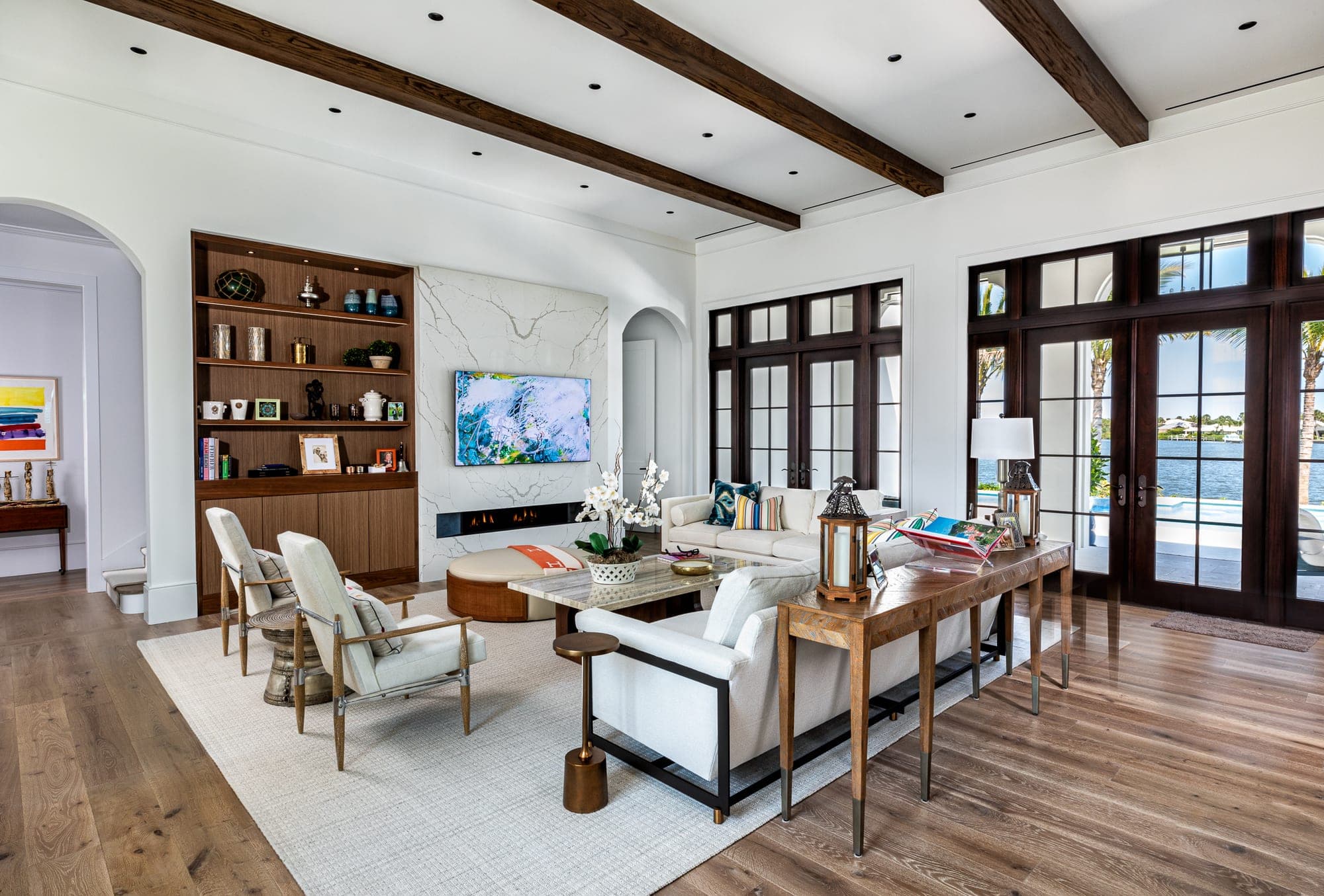 Living room beneath dark stained ceiling beams, with walnut built-in shelving, a stone slab fireplace wall and French doors to the river