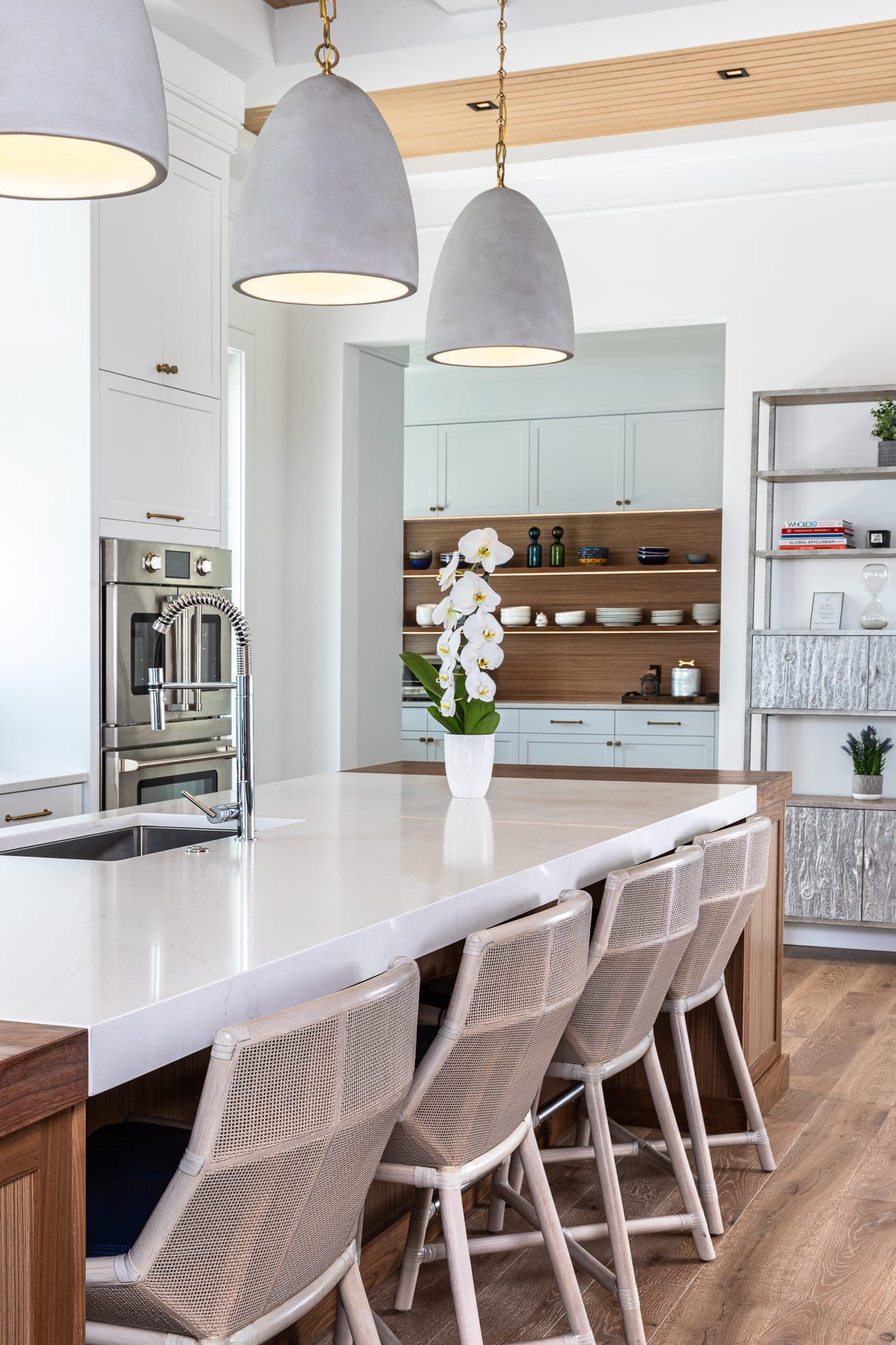 Kitchen with a white stone island on a walnut base, woven counter stools, three pendant lights and walnut open shelving beyond